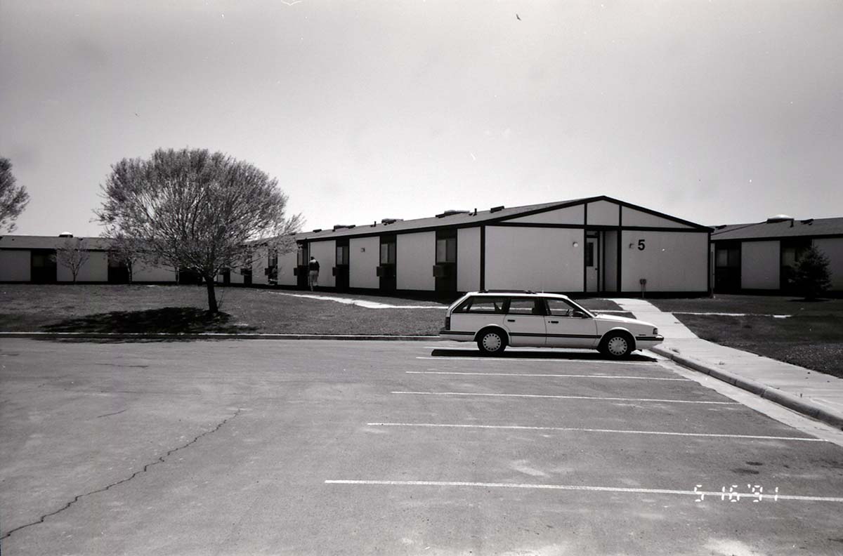 Barracks and parking area with man walking on sidewalk. Building number 5. [Image possibly for comparative housing study]