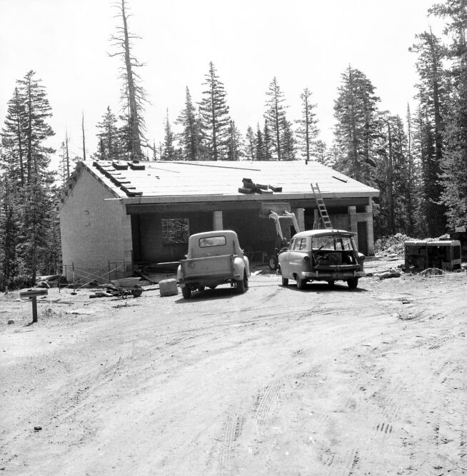 Utility and maintenance building under construction with stacks of shake shingles on roof. Trucks and equipment in front of building.
