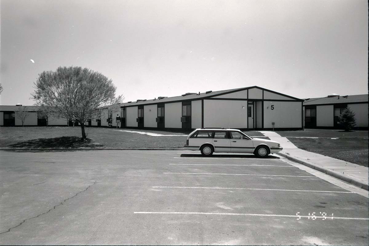 Barracks and parking area with man walking on sidewalk. Building number 5. [Image possibly for comparative housing study]
