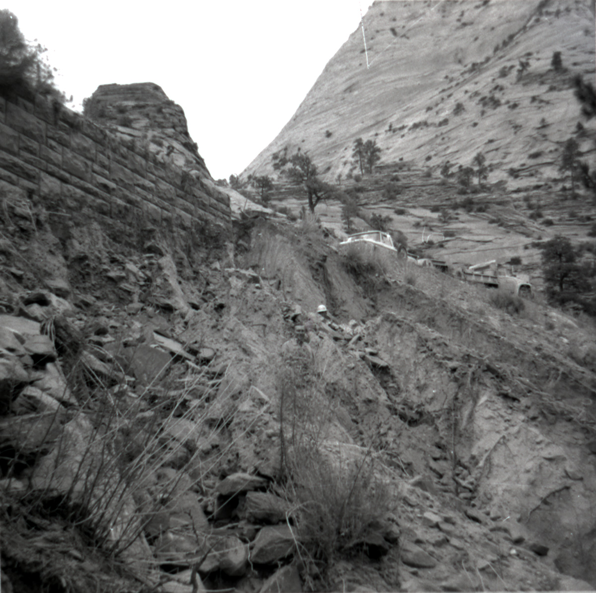 Men standing next to retaining wall in need of repair along East Rim road, trucks parked up on road.