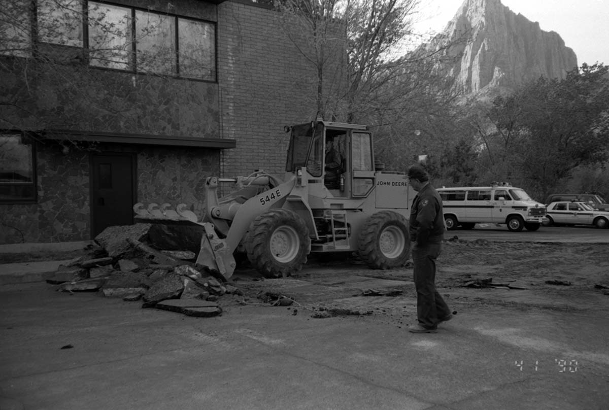 Worker operating an excavator machine while another worker looks on during construction of headquarters addition.