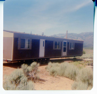 Single wide trailer home loaded onto truck trailer. Sage brush in foreground, mountains in background.